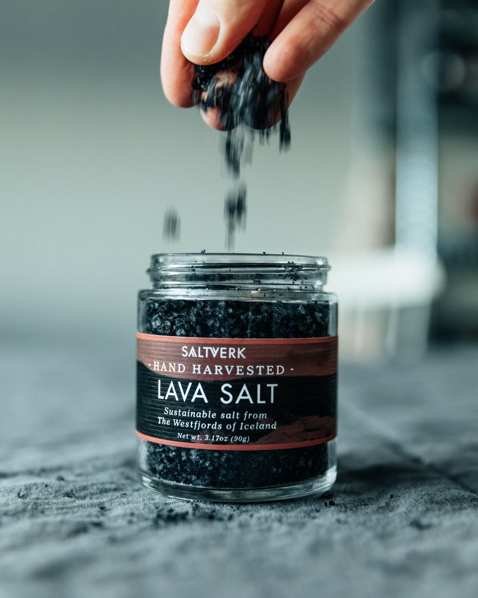 Jar of Saltverk lava salt with a hand sprinkling salt over it.